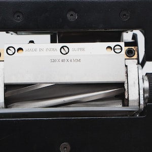 May include: Close-up of a metal blade assembly with the text "MADE IN INDIA" and "SUPER". The blade measures 120 x 40 x 4 mm. The assembly is set in a black metal frame.