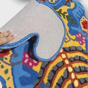 May include: A colorful, patterned rug with a blue border. The rug features a vibrant design with blue, yellow, red, and green floral and abstract elements. The underside of the rug is a light gray fabric.
