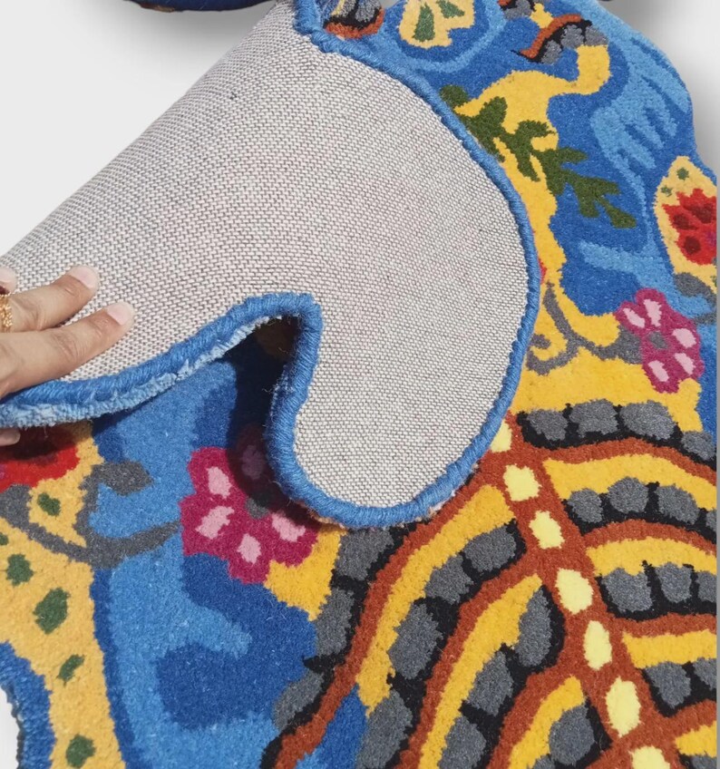 May include: A colorful, patterned rug with a blue border. The rug features a vibrant design with blue, yellow, red, and green floral and abstract elements. The underside of the rug is a light gray fabric.