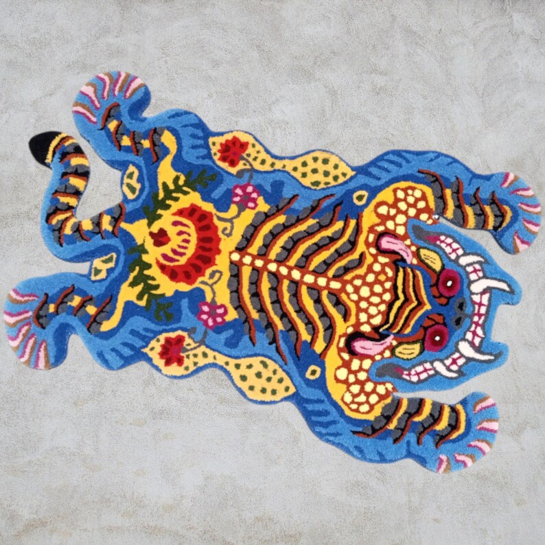 May include: A vibrant, hand-tufted rug in the shape of a tiger. The rug features a blue background with yellow, red, and gray details. The tiger's body has striped patterns, and floral accents add to the design. The rug is a decorative floor covering.