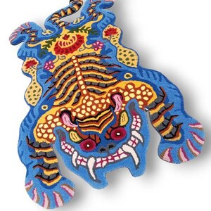 May include: A vibrant, intricately designed rug in the shape of a mythical creature. The rug features a blue base with detailed patterns in yellow, red, orange, and black. The creature has a fierce expression with sharp teeth and red eyes.