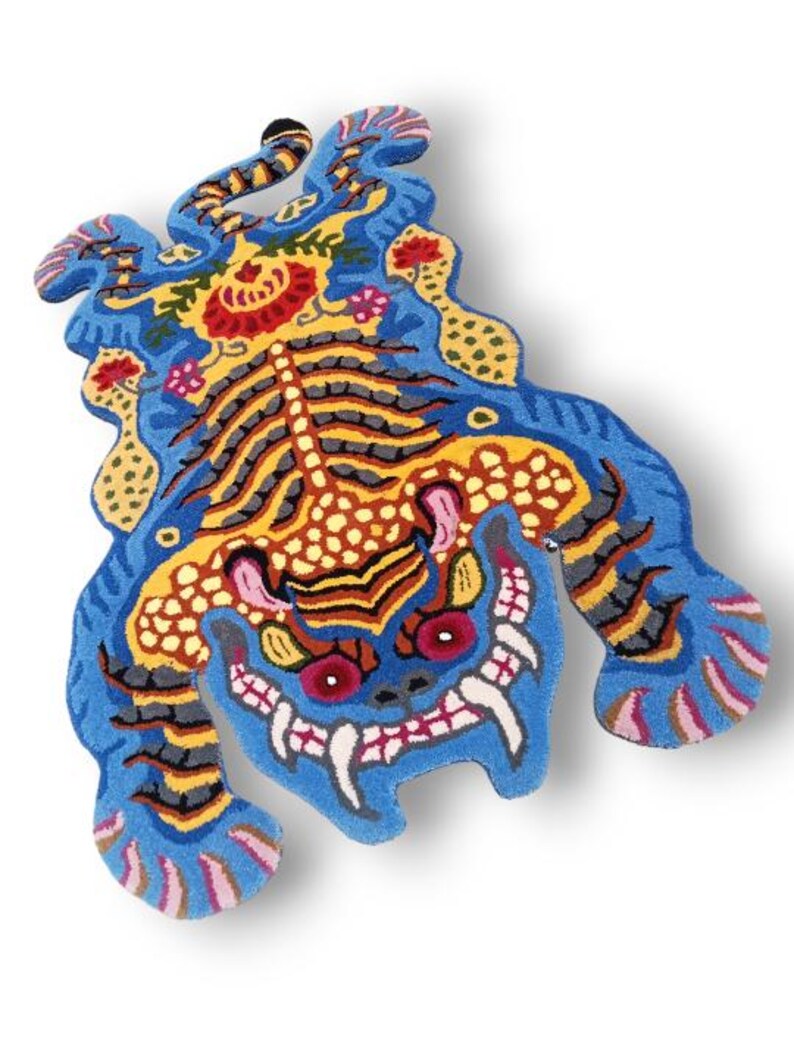 May include: A vibrant, intricately designed rug in the shape of a mythical creature. The rug features a blue base with detailed patterns in yellow, red, orange, and black. The creature has a fierce expression with sharp teeth and red eyes.