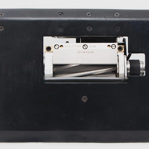 May include: A black industrial machine with a cylindrical handle on the left and a silver and black mechanism in the center. The mechanism has a rotating component and a small label with text. The machine is on a white background.