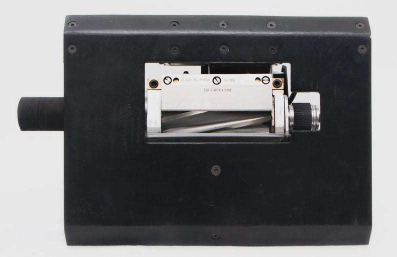 May include: A black industrial machine with a cylindrical handle on the left and a silver and black mechanism in the center. The mechanism has a rotating component and a small label with text. The machine is on a white background.