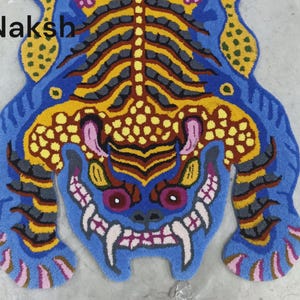 May include: A blue, yellow, and pink rug in the shape of a tiger. The rug has a detailed design with a tiger face and body. The word "Naksh" is in the upper left corner.