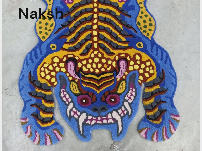 May include: A blue, yellow, and pink rug in the shape of a tiger. The rug has a detailed design with a tiger face and body. The word "Naksh" is in the upper left corner.