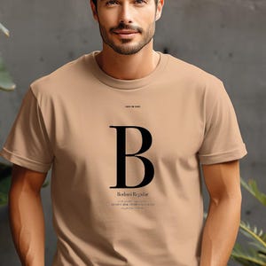 Könnte beinhalten: Ein beigefarbenes Kurzarm-T-Shirt mit einem großen schwarzen Buchstaben B in der Mitte der Brust. Über dem Buchstaben steht der Text "FONTS ON SHIRT" und darunter "Bodoni Regular", gefolgt von einer kleinen alphabetischen Zeichenfolge.