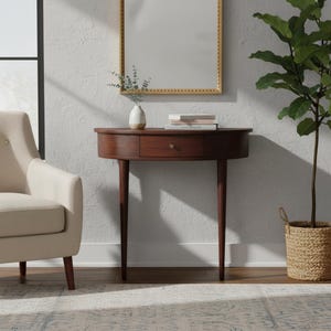 May include: A dark brown, semi-circular console table with a drawer and brass knob. The table has four slender, tapered legs. A small white vase with greenery, books, and a gold-framed mirror are on the table. A beige armchair and a potted plant are also in the room.