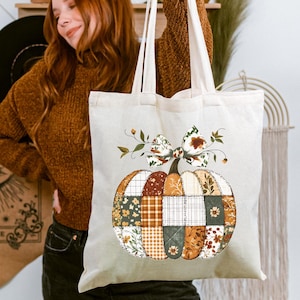 May include: Cream-colored tote bag featuring a patchwork pumpkin design. The pumpkin is composed of various fabric patterns in shades of orange, brown, green, and white, with a decorative bow and stem. The bag has sturdy handles.