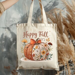 May include: Beige tote bag with a pumpkin design and the text "Happy Fall." The pumpkin is decorated with flowers and a bow. The bag is held by a person.