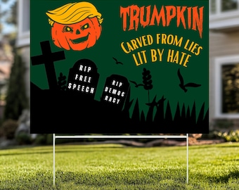 May include: A green yard sign with the word "Trumpkin" in orange, featuring a pumpkin with a blonde hairstyle. The sign includes the phrases "Carved from Lies Lit by Hate" and "RIP Free Speech, RIP Democracy."