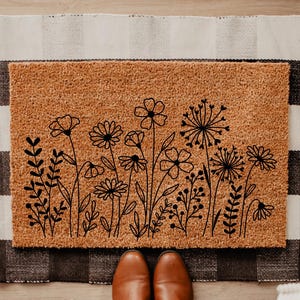 May include: A rectangular doormat made of natural coir fiber, featuring a black line drawing of various flowers and foliage. The mat is placed on a black and white checkered rug. Brown leather shoes are visible at the bottom of the image.