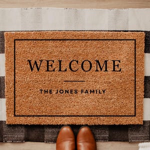 May include: A rectangular coir doormat with the word "WELCOME" in black, above "THE JONES FAMILY". The mat is framed in black and sits on a black and white checkered rug. Brown shoes are visible at the bottom.