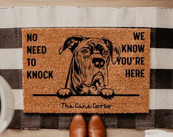 Könnte beinhalten: Rechteckige Fußmatte mit einer Cane Corso Hundeillustration und dem Text "NO NEED TO KNOCK WE KNOW YOU'RE HERE". Die Matte ist braun mit schwarzem Text und einer schwarzen Hundeillustration. Der Text "The Cane Corso" steht darunter.