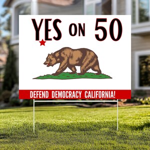 May include: A white yard sign with the text "YES ON 50" in red and black, featuring a brown bear and a red star. The bottom of the sign reads "DEFEND DEMOCRACY CALIFORNIA!" in white on a red banner. The sign is displayed on a green lawn.