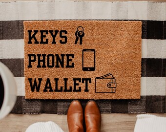 May include: A rectangular doormat with the words "KEYS PHONE WALLET" in black block letters. The mat has a brown, textured surface and includes line drawings of keys, a smartphone, and a wallet. The mat is on a black and white checkered rug.
