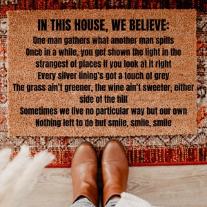 May include: A rectangular doormat with the text "IN THIS HOUSE, WE BELIEVE:" and a poem in black font. The mat is brown and sits on a patterned rug. Brown boots are visible at the bottom of the image.