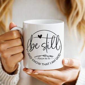 May include: White ceramic mug with the words "be still and know that I am God" in black script. The mug has a black heart and leaf design. The mug is held by a person.