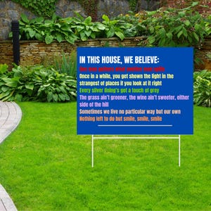 May include: A blue sign on a white metal frame, placed on a green lawn. The sign reads "IN THIS HOUSE, WE BELIEVE:" followed by a series of colorful lines of text. The sign is in front of a stone wall and lush greenery.