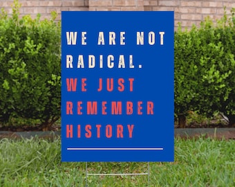 May include: A blue yard sign with white text that reads "WE ARE NOT RADICAL. WE JUST REMEMBER HISTORY." The words "WE JUST REMEMBER HISTORY" are in red. The sign is in a grassy yard with a brick wall and green bushes in the background.