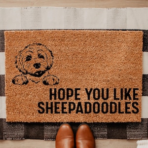 May include: A rectangular doormat with a brown coir surface features a black illustration of a sheepadoodle dog and the text "HOPE YOU LIKE SHEEPADOODLES". The mat is placed on a black and white checkered rug.