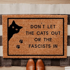 May include: A rectangular doormat with a black border and the text "DON'T LET THE CATS OUT OR THE FASCISTS IN." A black cat silhouette and paw prints are on the mat. The mat is on a black and white checkered rug.