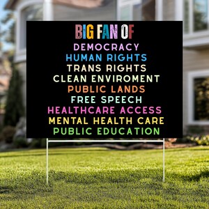 May include: A black yard sign with white metal stakes. The sign has the words "BIG FAN OF" in colorful letters at the top, followed by a list of values including "DEMOCRACY" and "PUBLIC EDUCATION". The sign is placed in a grassy yard.
