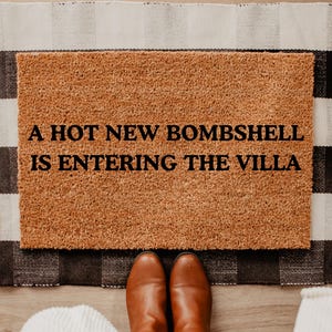 May include: A rectangular doormat with the text "A HOT NEW BOMBSHELL IS ENTERING THE VILLA" in black. The mat is brown and sits on a black and white checkered rug. Brown leather boots are visible.