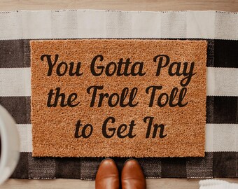 May include: A rectangular doormat with the text "You Gotta Pay the Troll Toll to Get In" in black lettering. The mat is brown and sits on a black and white checkered rug. Brown shoes are at the bottom.