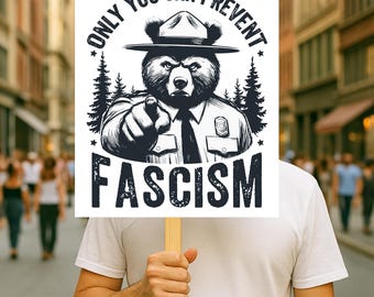 May include: A sign held by a person with the text "ONLY YOU CAN PREVENT FASCISM". The sign features a bear wearing a ranger hat and uniform, pointing forward. The background is a blurred street scene.