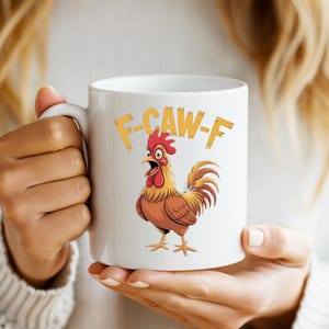 May include: White ceramic mug featuring a cartoon rooster with a red comb and brown feathers. The mug has the text "F-CAW-F" in yellow. The mug is held by a person.