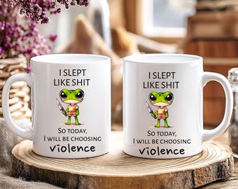 Könnte beinhalten: Zwei weiße Keramikbecher mit dem Text "I SLEPT LIKE SHIT" und "SO TODAY, I WILL BE CHOOSING violence". Jeder Becher zeigt einen Cartoon-Frosch, der ein Messer hält. Die Becher stehen auf einer Holzoberfläche.