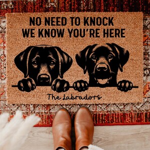 Può includere: Un zerbino rettangolare con la scritta "NO NEED TO KNOCK WE KNOW YOU'RE HERE" e due illustrazioni di labrador neri. Le parole "The Labradors" sono scritte sotto i cani. Il tappeto è marrone.