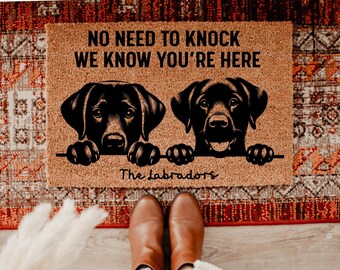 Può includere: Un zerbino rettangolare con la scritta "NO NEED TO KNOCK WE KNOW YOU'RE HERE" e due illustrazioni di labrador neri. Le parole "The Labradors" sono scritte sotto i cani. Il tappeto è marrone.