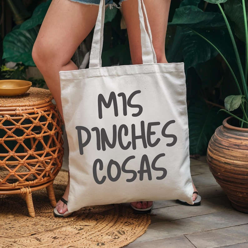 May include: Cream-colored tote bag with the phrase "MIS PINCHES COSAS" in dark gray lettering. The bag has two long handles and is held in front of a person. The background includes greenery and a woven side table.