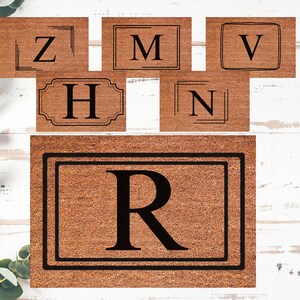 Può includere: Una collezione di zerbini marroni con bordi neri e grandi lettere in grassetto. Le lettere includono Z, M, V, H, N e R. I tappeti sono disposti su una superficie di legno bianco, con vegetazione sullo sfondo.