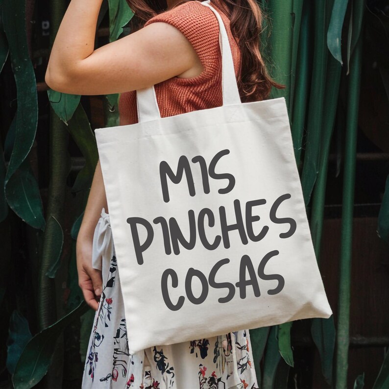 May include: Cream-colored tote bag with the phrase "MIS PINCHES COSAS" printed in dark gray. The bag has two white handles and is being held in front of a green leafy background.