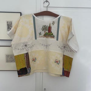 Op de afbeelding: Een top met korte mouwen gemaakt van een patchwork van crèmekleurige, gele en bedrukte stoffen. Het middenpaneel toont een geborduurd landschap met een toren en boom, geflankeerd door bloemmotieven en delen van gestructureerde, veelkleurige stof.