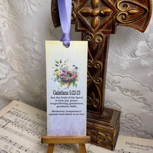 May include: A decorative bookmark with a floral design and the text "Galatians 5:22-23" is displayed on a small wooden easel. The bookmark has a purple ribbon attached. A bronze cross is in the background.
