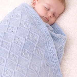 Puede incluir: Un bebé dormido envuelto en una manta de punto azul claro. La manta presenta un patrón de diamantes con detalles calados. El bebé descansa sobre una superficie blanca y esponjosa, creando una escena suave y reconfortante.