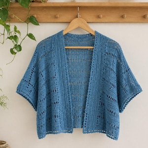 May include: A blue crochet cardigan with short sleeves hangs on a wooden hanger. The open-front sweater features a textured stitch pattern and a scalloped edge. The garment is displayed against a white wall with a wooden shelf and greenery.