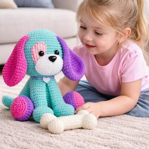 May include: A handmade crochet dog toy in teal, pink, and purple, with a white bone-shaped toy. The dog has large floppy ears and a heart-shaped patch. The toy is sitting on a light-coloured rug.
