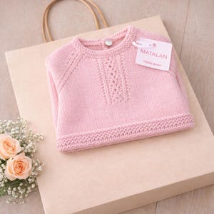 Puede incluir: Un suéter de bebé tejido de color rosa claro con un diseño texturizado en la parte delantera. El suéter tiene un dobladillo y puños acanalados, y un pequeño cierre de botón en la parte posterior del cuello. Una etiqueta dice "MATALAN PREM/BABY". El suéter está sobre una bolsa de papel marrón claro.