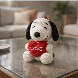 May include: A crocheted Snoopy plush toy, holding a red heart with the word "LOVE" embroidered in white. The toy has a white body, black ears, and a black nose. The plush is sitting on a glass surface.