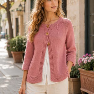May include: A dusty rose knit cardigan featuring three round wooden buttons, a round neckline, and three-quarter length sleeves with ribbed cuffs. The garment has a textured border along the front opening and hem, worn over a white top and light trousers.