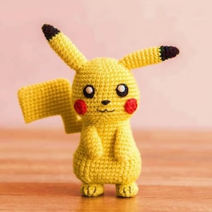 Puede incluir: Figura de Pikachu amarilla hecha a mano con ganchillo, con orejas con puntas negras, mejillas rojas y una pequeña cola. La figura está sobre una superficie de madera. La figura mide aproximadamente 15 cm de altura.