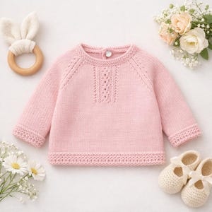 Puede incluir: Un suéter rosa claro para bebé con mangas largas y un patrón de punto decorativo. El suéter se muestra con un mordedor de orejas de conejo de madera, zapatos de bebé color crema y flores. El suéter tiene un cierre de botón en la espalda.