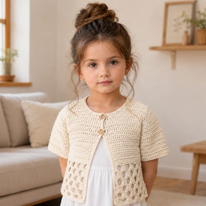 May include: A cream-colored, short-sleeved crochet cardigan featuring a round neckline, two small wooden buttons at the center front, and a decorative openwork stitch pattern along the lower section of the garment, worn over a plain white dress.
