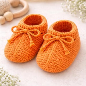 Op de afbeelding: Een paar oranje gehaakte babyslofjes met strikdetails. De slofjes zijn handgemaakt en hebben een gestructureerd steekpatroon. De schoentjes zijn ongeveer 10 cm lang, perfect voor een pasgeborene. De achtergrond bevat een houten speeltje en witte bloemen.