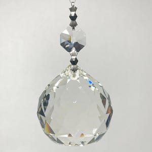 May include: A clear crystal ball suncatcher with multiple facets, suspended by a silver-colored chain. The crystal reflects light, creating a rainbow effect. The suncatcher is designed for decorative purposes.